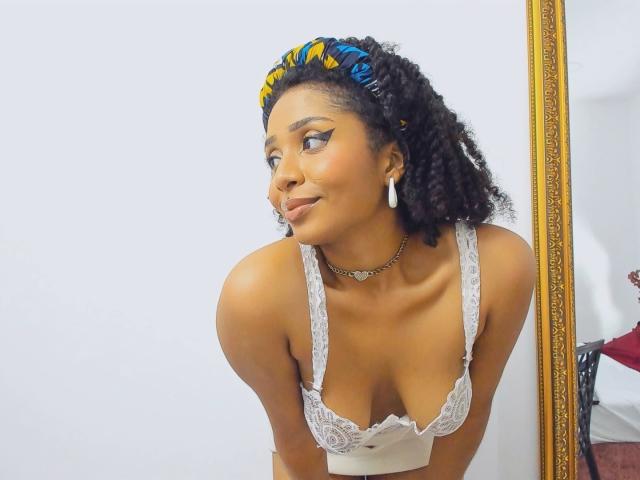MeganWilliams with dark curly hair, yellow and blue headband, white lace bra, showing her tits, smiling, earrings, necklace, leaning forward, reflection in ornate mirror.