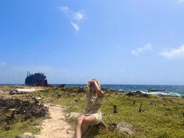 VictoriiaGrey with blonde hair, wearing a beige dress, sitting on a rock, hands covering her face, in a grassy, rocky coastal area with a rusted ship in the distance.
