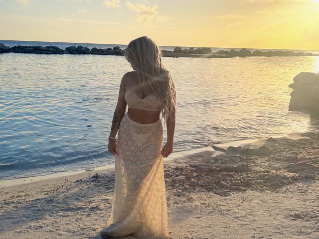 VictoriiaGrey with long blonde hair stands on a beach, facing the ocean. She's wearing a lace bra and skirt, showing her tan skin and small ass. Sunset in background.