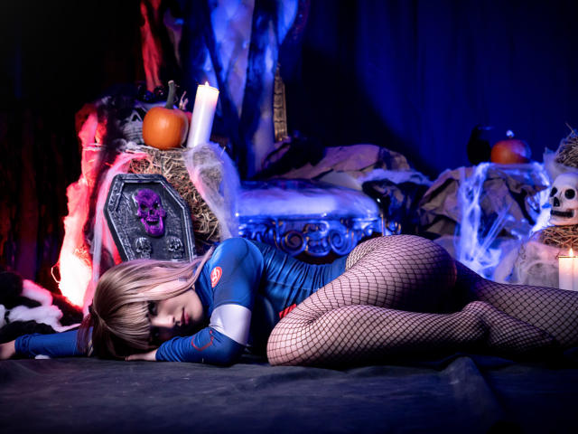 SamanthaVelvett, vestida con un traje azul, ajustado y futurista, con medias de red, está acostada de lado, mostrando sus glúteos y piernas, con una silla con forma de calavera y velas en el fondo..
