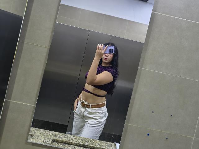 MarilynHills with dark wavy hair, purple crop top, white pants, holding phone in a bathroom mirror, showing her bare ass and brown belt.