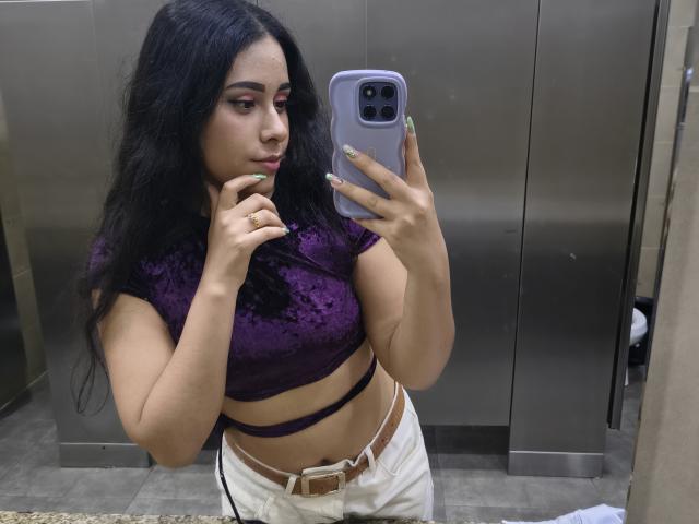 MarilynHills with long black hair, wearing a purple lace crop top, white pants, and a brown belt, taking a mirror selfie in an elevator, showing her tits.