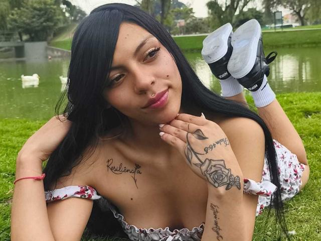 RoxannaMoretti with black hair, pink lipstick, tattoos on chest and hand, lying on grass, floral dress, white shoes, red bracelet, hands on chin, legs up, green park background.