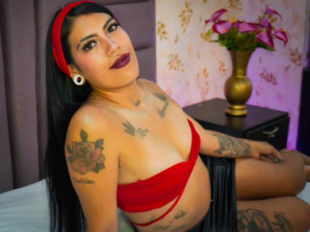 KymCorzo, avec de longs cheveux noirs, un bandeau rouge, un haut rouge sans bretelles, des tatouages, du rouge à lèvres foncé, assise sur un lit, regardant la caméra, montrant ses seins, une jupe noire plissée, un lit blanc, un vase..