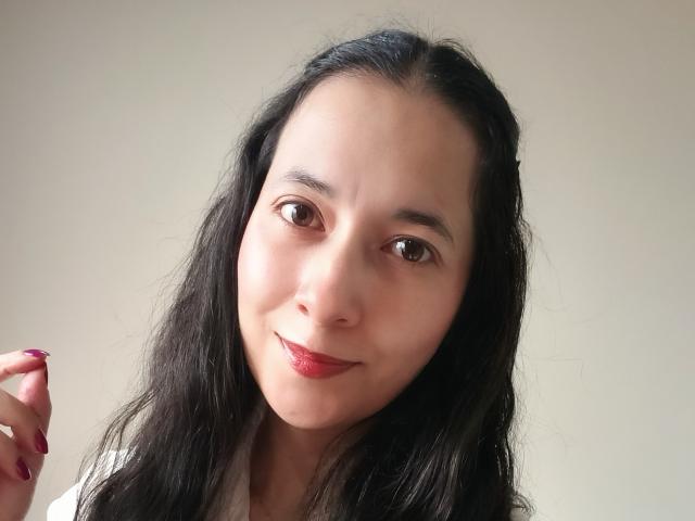 AnneStone has long black hair, brown eyes, and red lipstick, with a subtle smile. Her pink nail is visible, and she's wearing a white shirt.