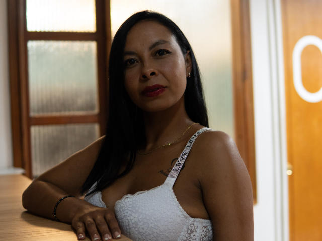 ClairBennet with dark hair, red lipstick, white lace bra, showing her tits, leaning on a wooden surface, gold necklace, bracelet, and tattoo on her right chest.