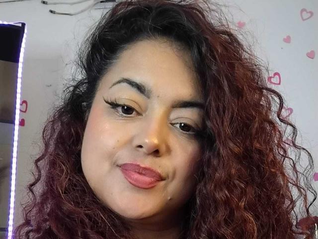 SophiLarusso has curly red-brown hair, full lips, and is pouting slightly. She has dark eyebrows, long eyelashes, and a small beauty mark under her right eye.