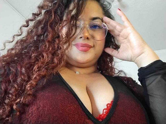 SophiLarusso with curly red-brown hair, glasses, and red lipstick, showing her large, lace-covered boobs in a sheer black and red top, touching her face.