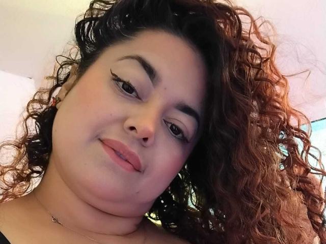 SophiLarusso has curly brown hair, light brown skin, and full lips with pink lipstick. She's looking slightly to the side with winged eyeliner and a subtle smile.