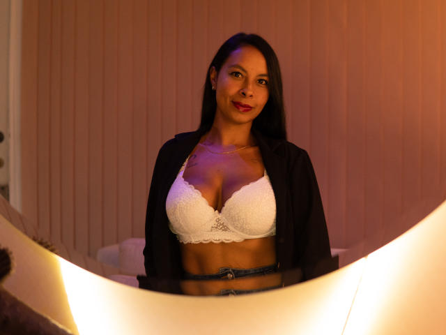 ClairBennet with medium brown skin, long black hair, wearing a white lace bra that shows her tits, black open shirt, and blue jeans, smiling in warm lighting.