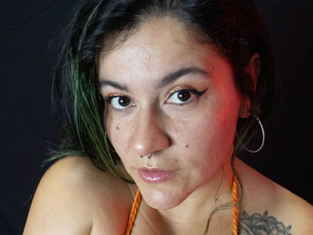 NaniSky with green-tinted hair, nose ring, and hoop earring, wearing an orange halter, has tattoos on her shoulder, and is looking at the camera with a pouty expression.