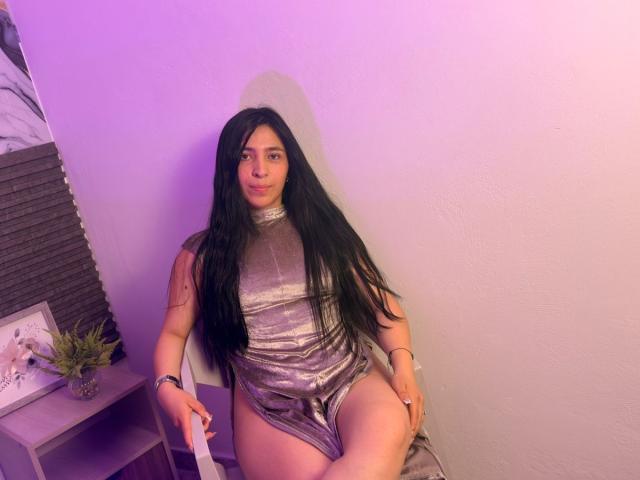 SamantaDiamont with long black hair, wearing a shiny silver dress, sitting on a chair, purple light, showing her ass, looking at camera, wrist bracelet, small potted plant on left.
