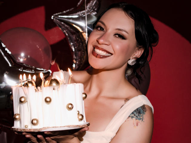 KatalinaJohnson with dark hair, smiling, holding a white cake with candles, gold balls, and wearing a dress, showing a tattoo on her right arm.