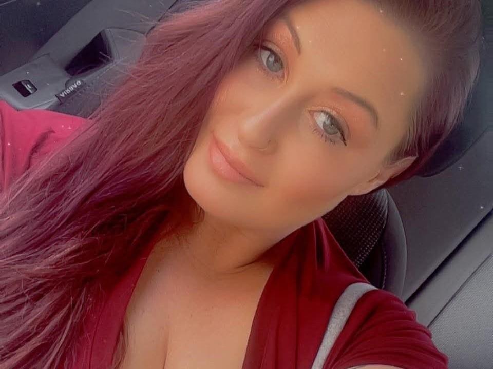 Con i capelli rossi, gli occhi verdi e un leggero sorriso, indossa una maglietta rossa con spalline grigie, che lascia intravedere un po' di scollatura. Illuminazione soffusa, trucco leggero..