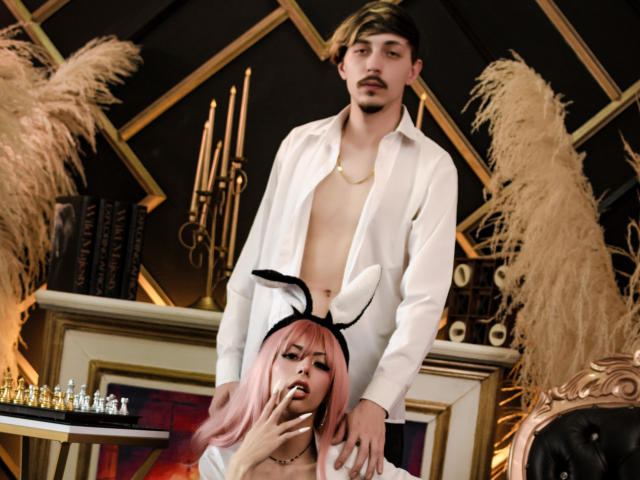 JunnaAndKurt with pink hair, bunny ears, and a cigarette, kneeling, wearing a white outfit, while Kurt stands behind, shirtless, with an open white shirt, and a gold necklace.