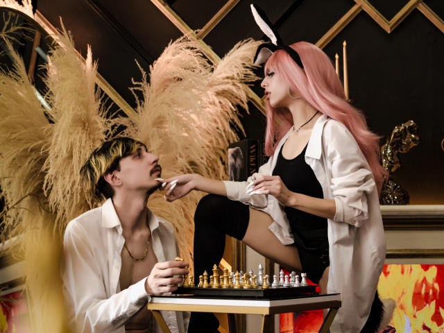 JunnaAndKurt with pink hair and white bunny ears, sitting on a table, wearing a black dress and white shirt, holding a cigarette, looking at Kurt with blond hair.