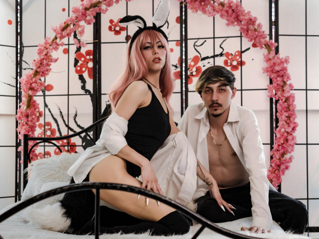 JunnaAndKurt with pink hair, black bunny ears, and black lingerie, sitting on a bed with Kurt, shirtless in a white shirt, black pants, and brown hair.