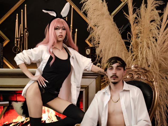JunnaAndKurt with pink hair, black bunny ears, white shirt, black tank, thigh-highs, standing, showing ass. Kurt, shirtless, white shirt, seated, looking at JunnaAndKurt.