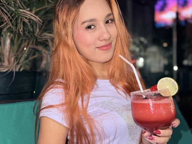 EmmaWat with long orange hair, holding a red cocktail with a straw, lime, and cherry, smiling at the camera, wearing a white shirt, in a dimly lit bar.