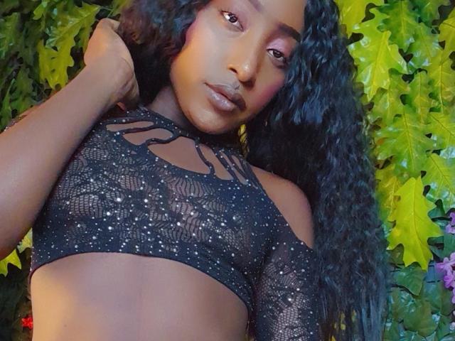 Jorinde with dark skin and curly black hair, wearing a black lace crop top with sparkles, showing her small, firm boobs. Leafy background. She's looking at the camera.