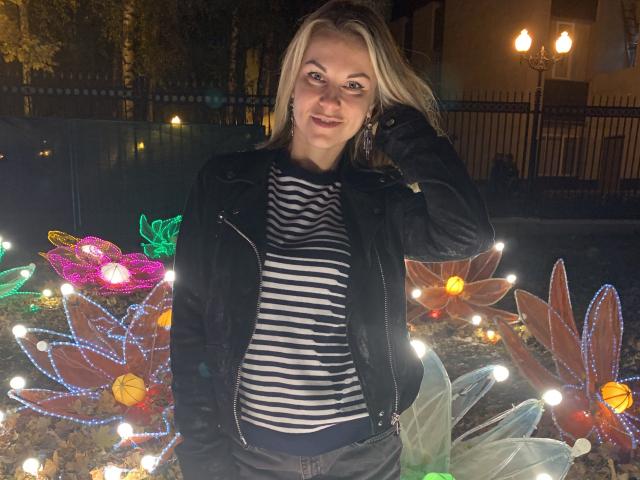 BelleLisaG with blonde hair, black jacket, striped shirt, smiling, hand in hair, standing in front of colorful Christmas flower lights, night, street lamp, dark fence, dark sky.