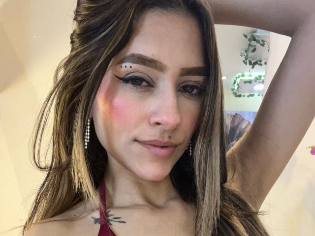 AfroditaWild mit langen, braunen Haaren, erhobener Arm, geflügeltem Eyeliner, kleinem Nasenpiercing, rosa Rouge, Tattoo an der rechten Schulter, Ohrringen, lächelnd, trägt ein rotes Halstuch-Top, Brust ist sichtbar..