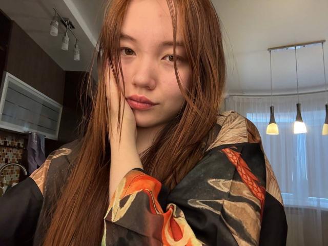 NereidaBottino with long red hair, pouty lips, and fair skin, wearing a floral black kimono, resting her hand on her cheek, in a softly lit room.