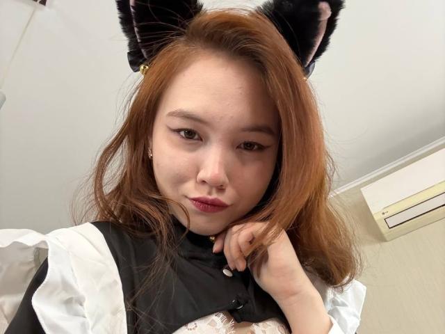 NereidaBottino with brown hair, wearing black cat ears and a black and white maid outfit, looking at the camera with a slight smile, her hand near her chin.