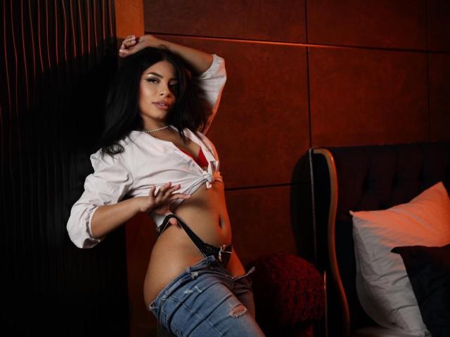 MissAvaa with long black hair, tied white shirt, revealing red bra, black thong, and unbuttoned jeans, leaning against a dark wall, looking at the camera with a sultry expression.