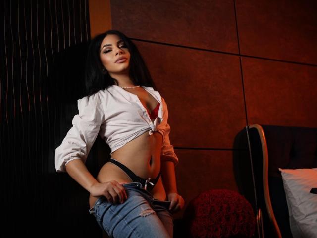 MissAvaa with black hair, tied white shirt, red bra, black thong, and ripped jeans, standing in a dimly lit room, hand on her jeans, showing her toned, curvy body.