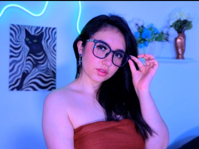 SusanMonroy with black hair, glasses, and pink lipstick, adjusting her glasses, wearing an orange strapless top, showing her tits. Blue neon light in the background.