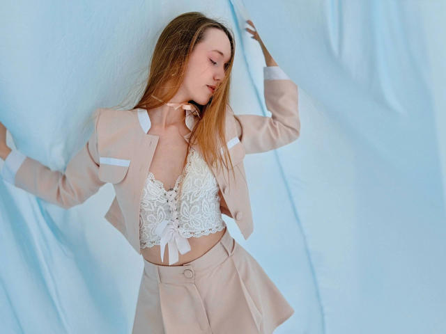 SunshineGirlX-hot in a beige suit with white lace top, showing her tits, blonde hair, pink choker, standing against a blue fabric background, arms raised, looking to the side.