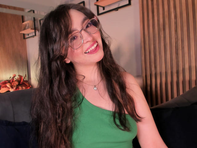 EvaMcAllister with long, wavy brown hair, green one-shoulder top, and glasses, smiling at the camera, showing her cute face and small tits. She's wearing a heart necklace.
