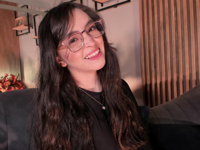 Eva McAllister, con cabello negro largo y ondulado, usando gafas y una camisa negra, sonriendo con labios rosados, mostrando dientes blancos pequeños y un collar de plata. Sus senos no son visibles..