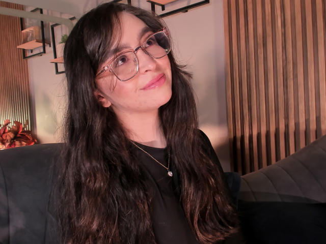 Eva McAllister, con cabello largo, oscuro y ondulado, gafas rosas, lápiz labial rosa, camisa negra, collar, sonriendo, sentada en un sofá de cuero negro, con una pared de paneles de madera en el fondo..