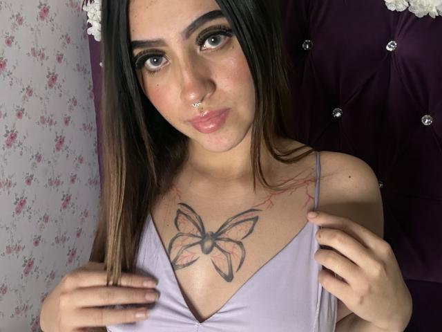 LunaaBlossom with brown hair, nose ring, and butterfly tattoo on chest, wearing a purple top, looking at camera with hands on straps. She has a cute, slightly pouty face.