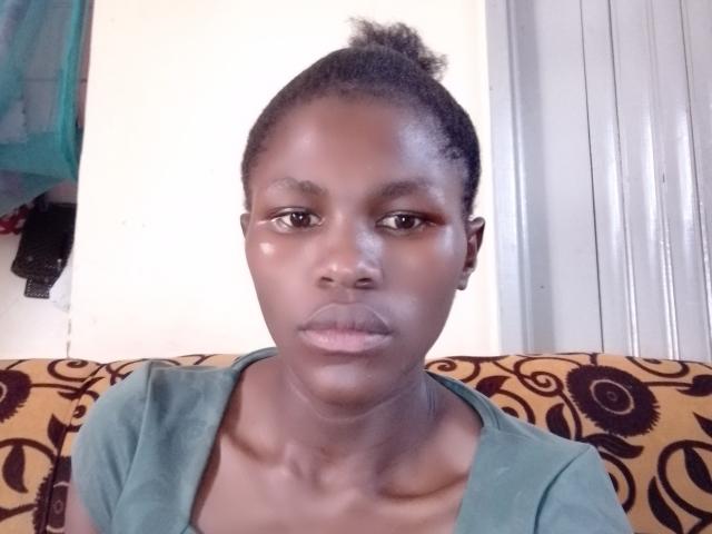 Valex69 a la peau foncée, de petits yeux marron et un chignon. Elle porte une chemise verte, et son derrière est légèrement visible sous la chemise. Elle regarde directement l'appareil photo..