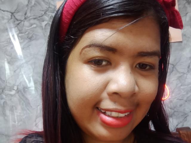 O rosto de MariahSquirting69, cabelo preto e liso, faixa vermelha, olhos castanhos, sorriso discreto, batom vermelho, nariz visível, pele lisa, parede de mármore ao fundo, luz suave, selfie casual..