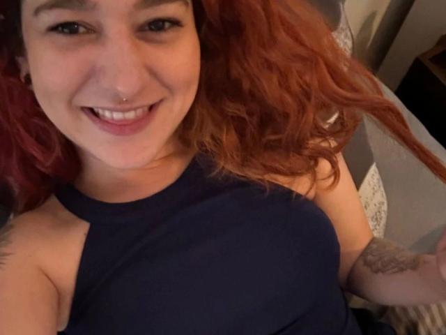 Mulher com cabelo ruivo, sorrindo, vestindo um top de alça azul marinho, com piercing no nariz, tatuagem no braço, deitada, mostrando os seios, e rosto bonito em uma selfie..