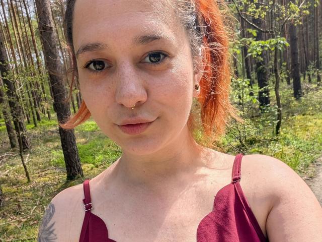 Bad-X-Tamy-hot with orange ponytail, nose ring, small tits in red tank top, forest background, freckles, subtle smile, light skin, tattoo on right shoulder.