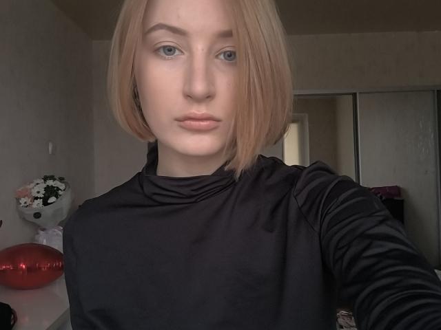 Edith Clantz, mit einer kurzen, blonden Bobfrisur, trägt eine schwarze Seidentop und hat blaue Augen und volle Lippen. Sie blickt direkt in die Kamera, und das Foto wurde in einem Raum mit neutralen Farben aufgenommen..