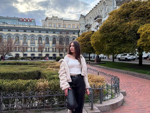 BiancaMassimo stands in a park, wearing a white top, black pants, and a fluffy beige jacket, with long brown hair and a cute ass, near ornate greenery.