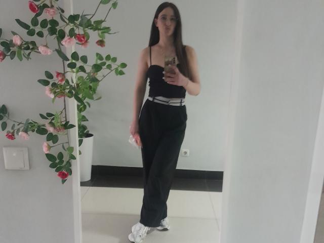 Omella with long dark hair, wearing a black strapless top and black pants, holding a phone, standing in a white room with pink roses, white sneakers.