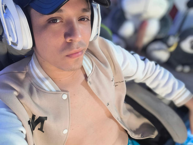 LukaForge with medium skin tone, wearing a beige varsity jacket, no shirt underneath, blue cap, white headphones, looking down with a neutral expression. Blurred background.