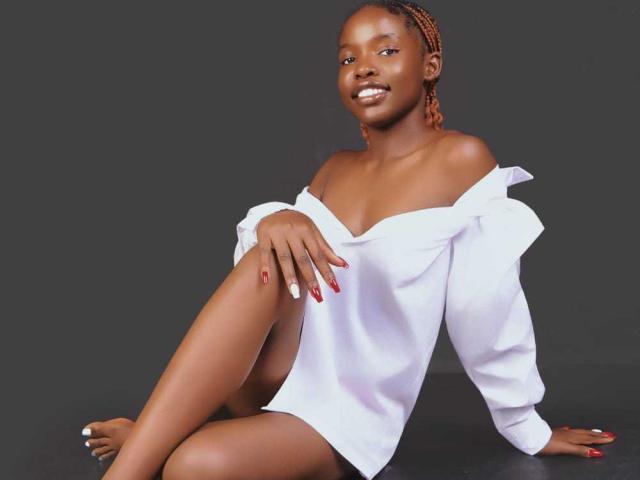KinkyAria with dark skin, small tits, no ass visible, wearing a white off-shoulder shirt, sitting, smiling, red and white nails, braided hair, dark background, sexy.