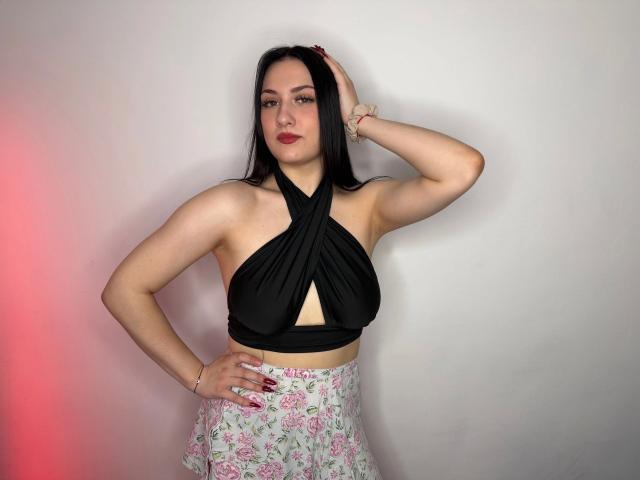 Darkling-hot with black halter top showing tits, floral skirt, hand on head, pink nail polish, red lipstick, white wall, red light, and bracelet, standing confidently.