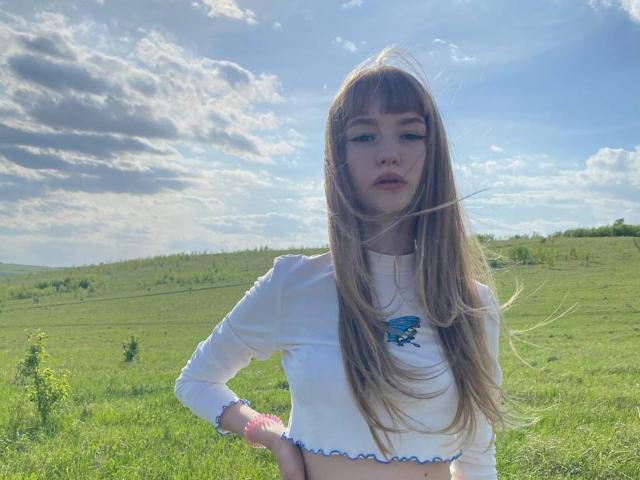 MargeryDoster with long brown hair, white crop top, and blue necklace, standing in a grassy field, sunlight, and cloudy sky, hands on hips, looking serious, tits visible.