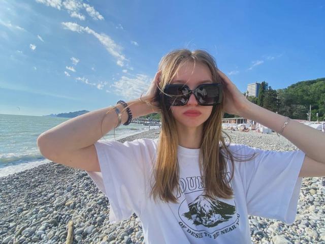 MargeryDoster with hands in her blonde hair, wearing black sunglasses, white t-shirt, and bracelets, standing on a rocky beach with a blue sky and ocean.