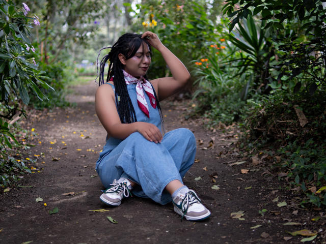 BeckyHuston seduta su un sentiero di terra, indossa una tuta in denim blu, scarpe da ginnastica bianche con lacci neri, una sciarpa rossa e bianca e trecce nere, e sta toccando la sua testa..