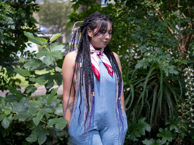 BeckyHuston with long black and purple braids, wearing blue denim overalls and a pink bandana, standing in front of lush green plants, smiling slightly, hands in pockets.